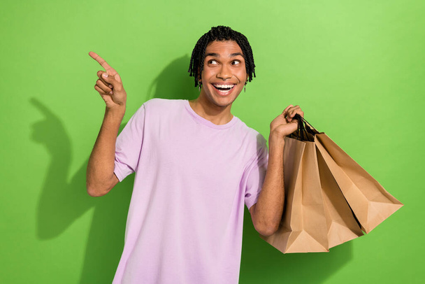 Photo of nice handsome guy toothy smile hold shopping bags look indicate finger empty space isolated on green color background. - Foto, Imagem