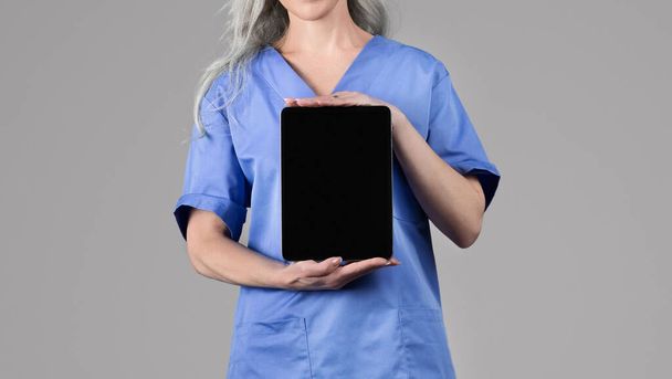 Mavi üniformalı tanınmayan hemşire kadın dijital tablet gösteriyor, gri stüdyo arka planında boş siyah ekran reklamcılık uygulaması gösteriyor. Kopyalama alanı olan Panorama - Fotoğraf, Görsel