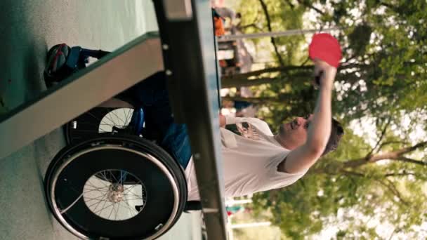 pionowe wideo Mężczyzna na wózku inwalidzkim gra w tenisa stołowego w parku miejskim emocjonalnie ćwiczy swoje ujęcia za pomocą rakiety do ping ponga - Materiał filmowy, wideo