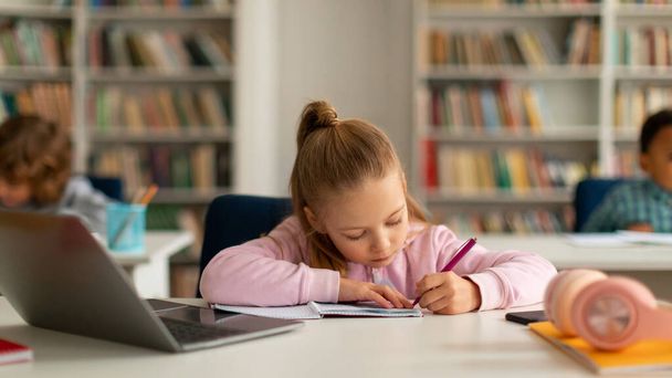 Skup się kaukaska uczennica pisanie w copybook podczas korzystania z laptopa komputera, siedzi przy biurku z kolegami na tle w szkole podstawowej, panorama - Zdjęcie, obraz
