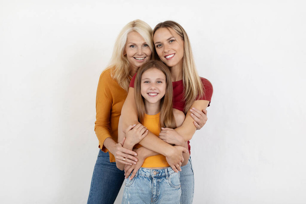 Love, family, affection concept. Beautiful preteen girl, millennial woman and senior lady bonding posing on white background. Happy three generations mother, daughter and grandmother embracing - Photo, Image
