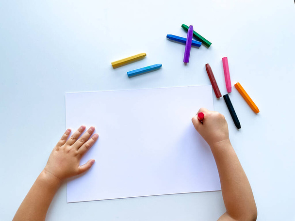 Kinderhände malen mit farbigen Wachsmalstiften auf ein weißes Blatt Papier. Draufsicht auf ein leeres Blatt. Hohe Qualität - Foto, Bild