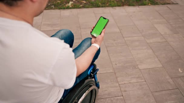 Un uomo su una sedia a rotelle tiene un telefono con uno schermo verde aperto in vista del parco cittadino da dietro - Filmati, video