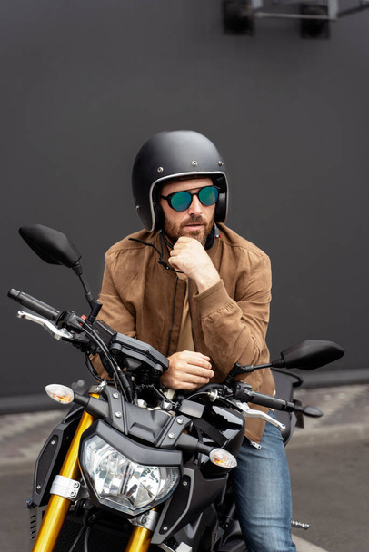 Βίαιος γενειοφόρος, ποδηλάτης φορώντας κράνος κάθεται σε σπορ μοτοσικλέτα στο δρόμο. Όμορφο επιτυχημένο μοντέλο μόδας φορώντας κομψό δερμάτινο μπουφάν ποζάρουν για φωτογραφίες σε μοτοσικλέτα  - Φωτογραφία, εικόνα