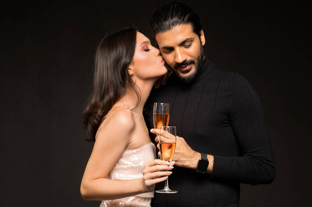 Loving multicultural young couple wearing beautiful outfits drinking champagne on black background, pretty brunette woman kissing indian guy boyfriend, lovers celebrating New Year, Christmas - Photo, Image