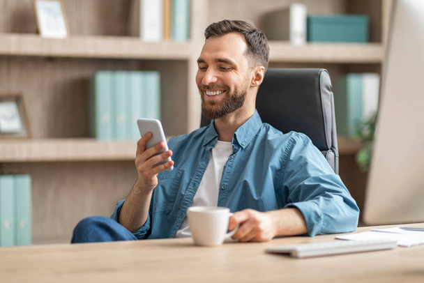 ナイスアプリ。 スマートフォンでリラックスし,職場でコーヒーを飲む若いビジネスマン,デスクで座っているカジュアルな服の陽気なミレニアル男性,ホットドリンクと携帯電話でメッセージング - 写真・画像