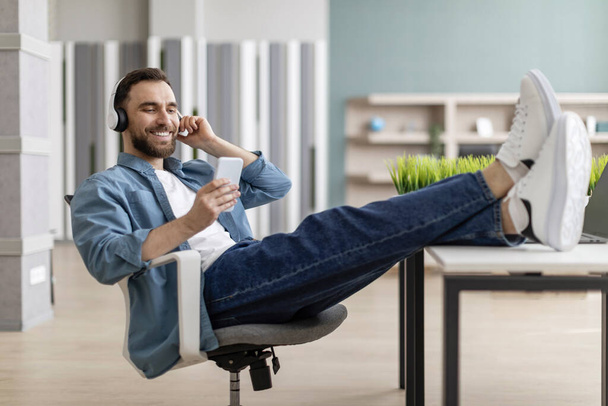 Streaming App. Smiling Young Man With Headphones And Smartphone Listening Music In Office, Handsome Millennial Guy Relaxing With Legs On Table, Enjoying Favorite Playlist, Resting At Workplace - Foto, Bild
