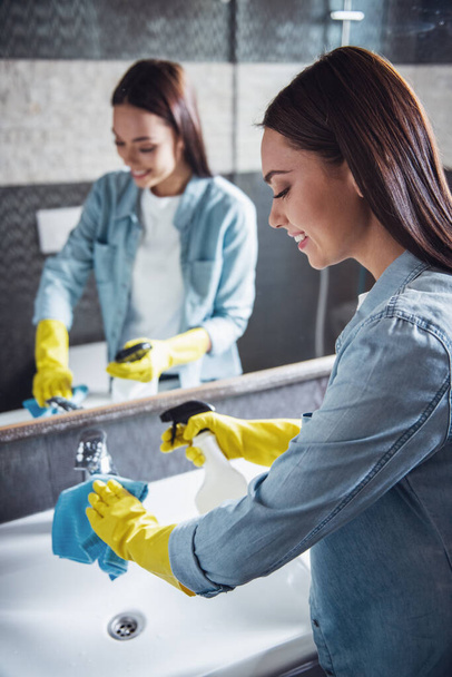 Hermosa mujer joven está utilizando un plumero y un detergente y sonriendo mientras limpia fregadero y grifo en el baño
 - Foto, Imagen
