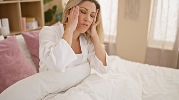 Junge blonde Frau leidet unter Kopfschmerzen, wenn sie im Schlafzimmer auf dem Bett sitzt - Filmmaterial, Video