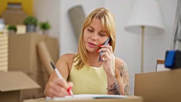 Mujer rubia joven hablando en un smartphone escribiendo en un cuaderno en un nuevo hogar - Imágenes, Vídeo