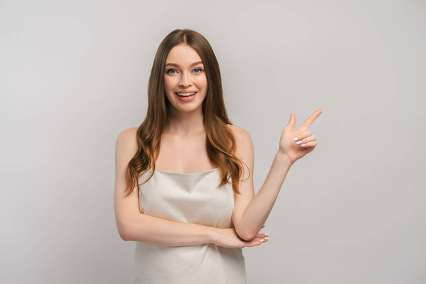Beautiful smiling woman with long beautiful hair wearing nightgown or dress pointing with finger on copy space isolated on gray background. Attractive girl posing in studio looking at camera  - Photo, Image