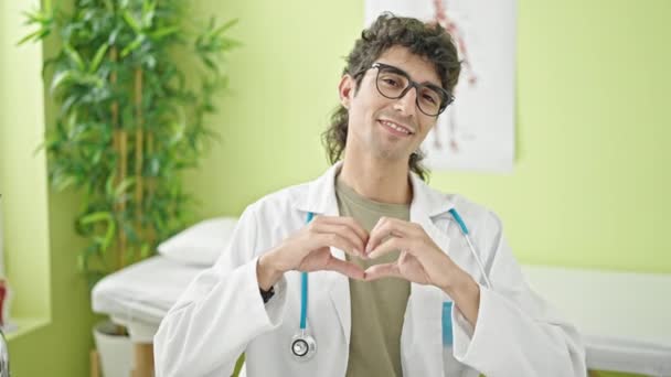 Joven médico hispano sonriendo haciendo gesto de corazón con las manos en la clínica - Metraje, vídeo