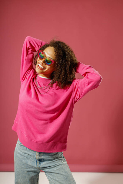 Улыбающаяся женщина в свитере касается волос и смотрит в сторону стоя на розовом фоне студии - Фото, изображение