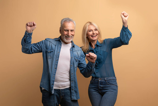 ベージュスタジオのバックグラウンドで成功を祝うカジュアルな服を着た幸せな退職夫婦と女性,手を挙げて笑顔を上げる拳をクレンチ - 写真・画像