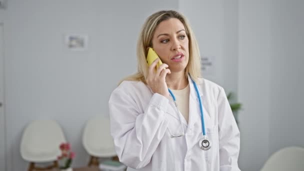 Mladá blondýnka žena lékař mluví na smartphone na klinice čekárna - Záběry, video