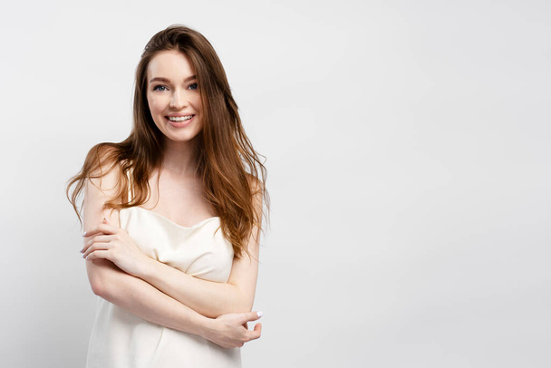 Portrait of young smiling woman with crossed arms wearing nightgown with long beautiful hair looking at camera isolated on gray background copy space. Attractive female, fashion model posing in studio - Photo, Image