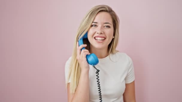 Młoda blondynka uśmiecha się pewnie rozmawiając przez telefon na odosobnionym różowym tle - Materiał filmowy, wideo