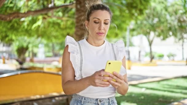 Mujer rubia joven usando smartphone celebrando en el parque - Imágenes, Vídeo