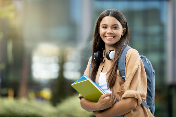 Happy beautiful long-haired hispanic young lady in casual outfit with wireless headphones and backpck going to college, posing at university campus, holding exercise books, copy space - Photo, Image
