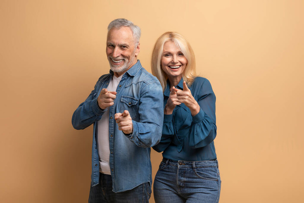 Уверенный холодный привлекательный кавказский пожилой мужчина и женщина в повседневной джинсовой одежде жестом указывая на камеру и улыбаясь, изолированные на бежевом фоне. Успешные пенсионеры - Фото, изображение
