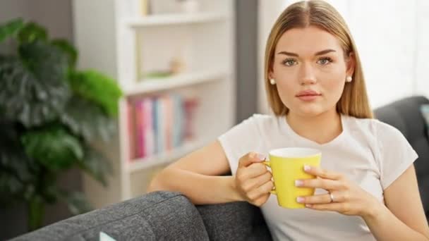 Νεαρή ξανθιά γυναίκα που πίνει καφέ καθισμένη στον καναπέ στο σπίτι - Πλάνα, βίντεο