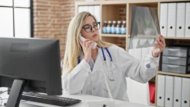Nuori blondi nainen lääkäri puhuu puhelimessa tilalla röntgenkuva klinikalla - Materiaali, video