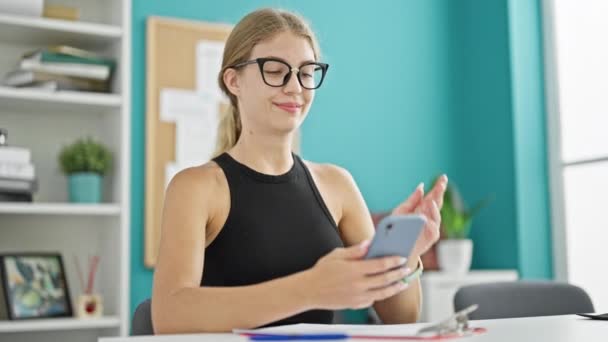 Młoda blondynka biznes pracownik za pomocą smartfona z gestem zwycięzca w biurze - Materiał filmowy, wideo