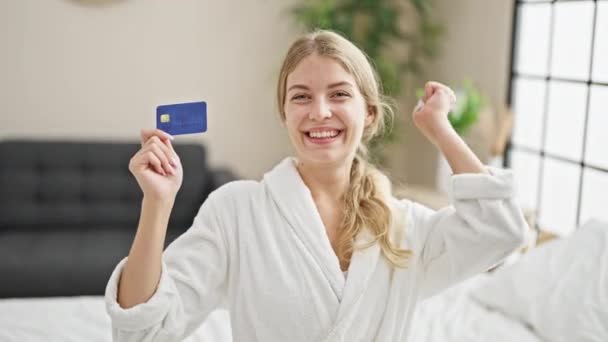 Fiatal szőke nő visel fürdőköpeny ül az ágyon kezében hitelkártya ünneplő a hálószobában - Felvétel, videó