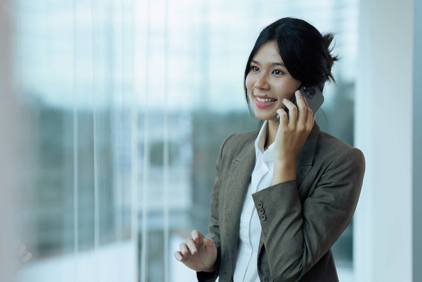 Een lachende zakenvrouw die door het raam kijkt terwijl ze op kantoor met een mobiele telefoon praat. Kopieerruimte. - Foto, afbeelding