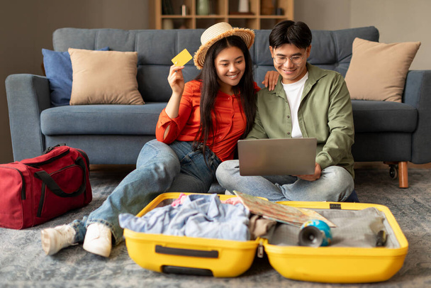 Happy japanese spouses using laptop and booking hotel online with credit card, holding passports with tickets at home. Travelers couple sitting with suitcases making vacation tour reservation - Photo, Image