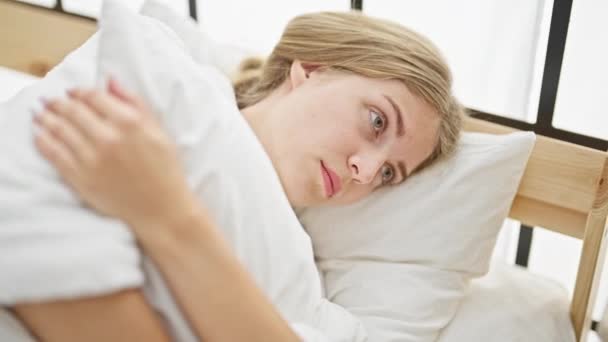 ベッドルームで深刻な顔で枕を抱擁するバスローブを着用している若い金髪の女性 - 映像、動画