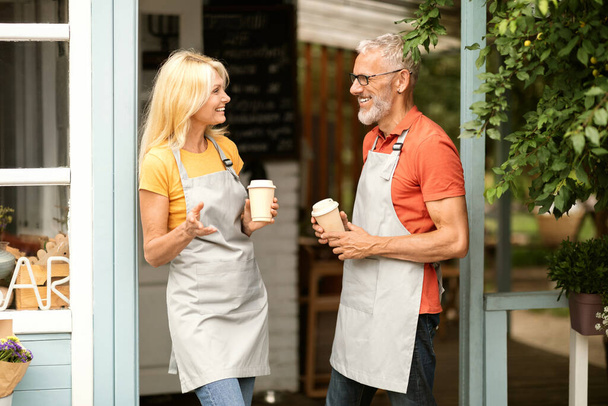 Happy Mature Barista Couple Chatting Outdoors While Standing At Coffee Shop Terrace, Smiling Older Man And Woman Wearing Aprons Talking And Having Drink, Enjoying Break At Work, Free Space - Photo, Image