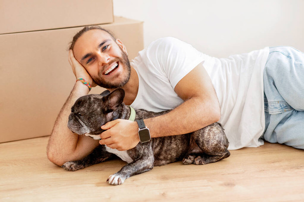 Szczęśliwy stylowy millenium facet hipster leżący na podłodze w pustym domu wśród kartonów z jego rzeczy, pieszcząc swojego psa w nowym mieszkaniu, korzystając z nowego domu. Wynajem, przeprowadzka, przeprowadzka - Zdjęcie, obraz