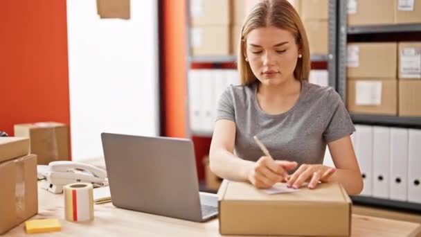 若い金髪女性 eコマースビジネスワーカー オフィスでパッケージにノートパソコンの書き込みを使用して - 映像、動画