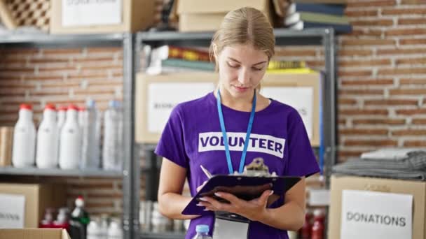 Młoda blondynka wolontariuszka uśmiecha się pewnie pisząc na liście kontrolnej w centrum charytatywnym - Materiał filmowy, wideo