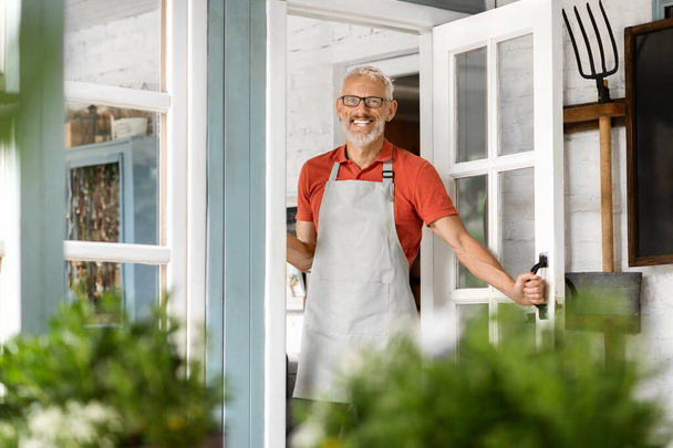 Красивый пожилой человек в фартуке и приветствуя в загородном доме, счастливый старший джентльмен позирует на террасе, современный мужчина фермер смотрит и улыбается в камеру, копирует пространство - Фото, изображение