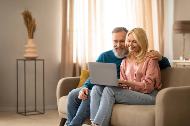 Счастливая пожилая европейская пара видеозвонит через ноутбук, проводит онлайн-встречу с семьей, сидит дома на диване в гостиной. Пенсионеры веб-серфинг на ПК, просмотр видео. Свободное пространство - Фото, изображение