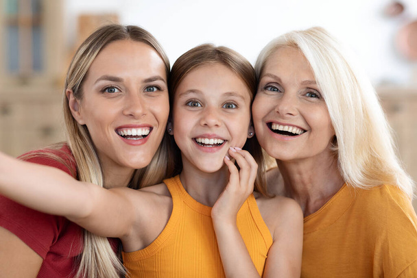 Gelukkig aantrekkelijk blond duizendjarige moeder, tiener dochter en grootmoeder nemen groep selfie thuis, drie familie generaties genieten van tijd samen, het nemen van foto 's, closeup - Foto, afbeelding