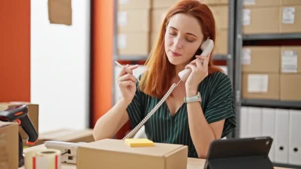 Νεαρή κοκκινομάλλα επιχειρηματίας ηλεκτρονικού εμπορίου που μιλάει στο τηλέφωνο γράφοντας σε χαρτί υπενθύμισης στο γραφείο - Πλάνα, βίντεο