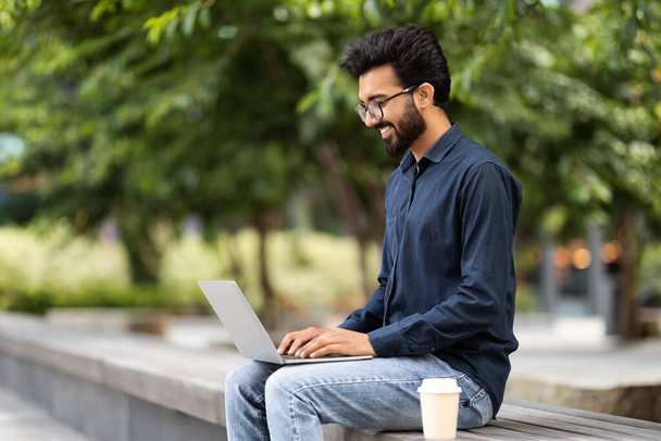 Positive handsome bearded eastern man remote employee working at public park, sitting on concrete bench, typing on laptop computer keyboard, drinking takeaway coffee, copy space - Photo, Image