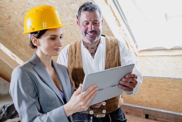 female architect wearing a hard heat and a carpenter on a construction site interior looking at a tablet computer showing blueprints of the project - Foto, imagen