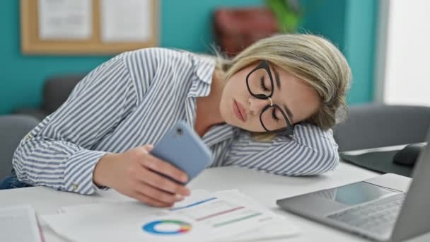 Nuori blondi nainen liiketoiminnan nainen työskentelee tylsää ja väsynyt käyttämällä älypuhelimen toimistossa - Materiaali, video