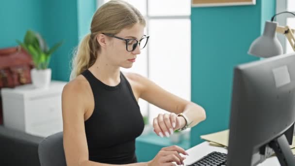 Νεαρή ξανθιά εργαζόμενη επιχείρηση χρησιμοποιώντας υπολογιστή αναζητούν ρολόι κουρασμένος στο γραφείο - Πλάνα, βίντεο