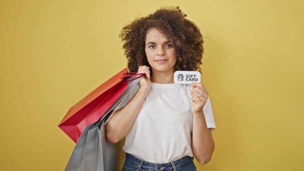 ショッピングバッグとギフトカードを保持している若い美しいヒスパニックの女性は,孤立した黄色の背景に微笑みます - 映像、動画