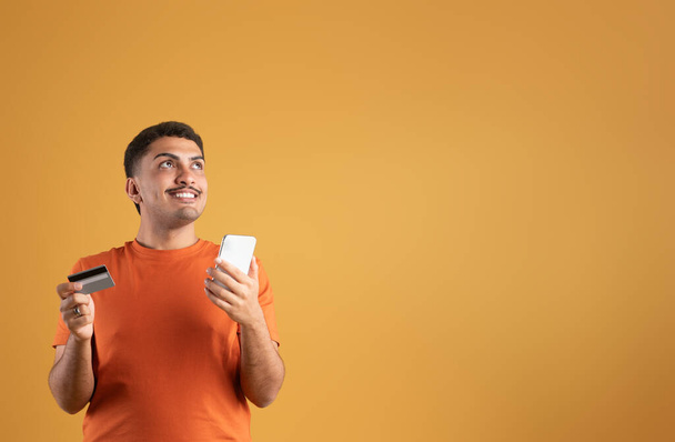 Χαρούμενος Βραζιλιάνος που κρατάει πιστωτική κάρτα και κινητό για online αγορές, κοιτάζοντας το χώρο των αντιγράφων στο φόντο του πορτοκαλί στούντιο. Ηλεκτρονικό χρήμα, διαχείριση διαδικτυακών οικονομικών - Φωτογραφία, εικόνα