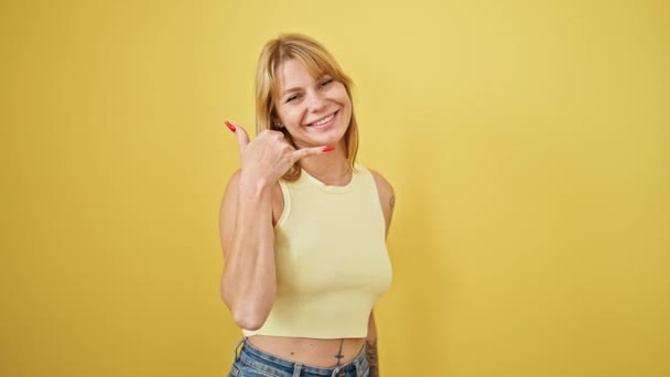 Junge blonde Frau lächelt selbstbewusst und macht Telefongeste mit Hand über isoliertem gelben Hintergrund - Filmmaterial, Video