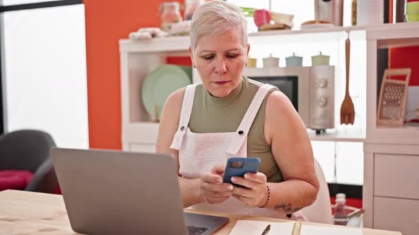 Mulher de cabelos grisalhos de meia idade usando smartphone e laptop na sala de jantar - Filmagem, Vídeo