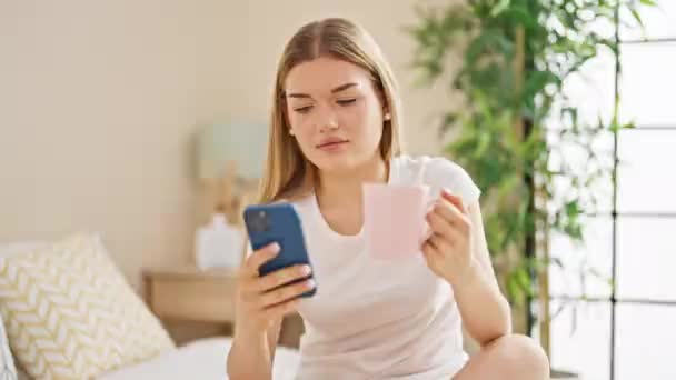 Mujer rubia joven usando smartphone bebiendo café en el dormitorio - Metraje, vídeo