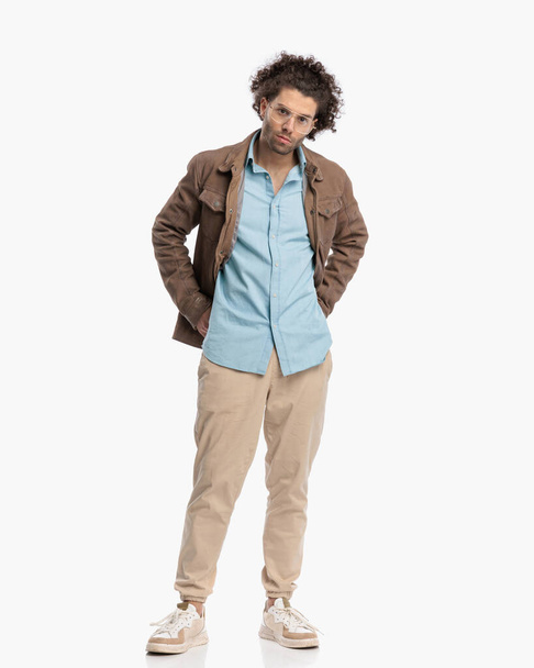 young casul man with curly hair holding hands in back pockets and posing while standing on white background - Foto, imagen