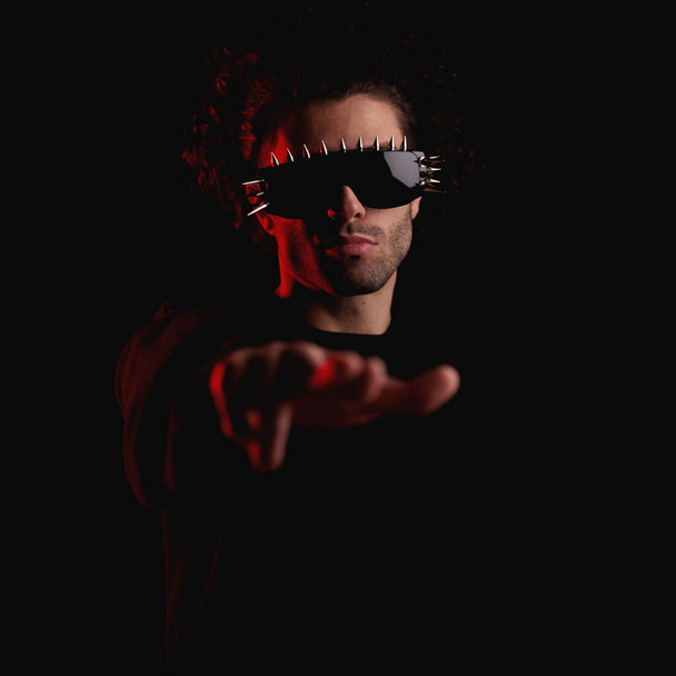 Kıvırcık saçlı, güneş gözlüğü takan ve siyah arka planda kırmızı ışıklarla parmakla işaret eden havalı bir adam. - Fotoğraf, Görsel